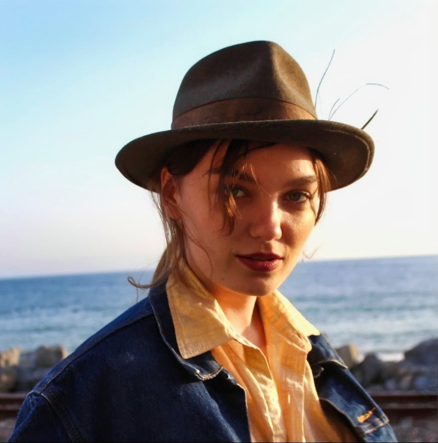 color photograph of a young woman looking at camera wearing a hat with the ocean in the background