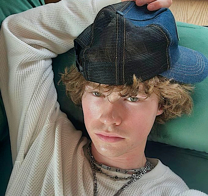 A color photograph of a young man with curly light brown hair lying down on a couch, wearing a dark mesh baseball cap backward and a white textured long-sleeve shirt looking directly at the camera in soft natural lighting.