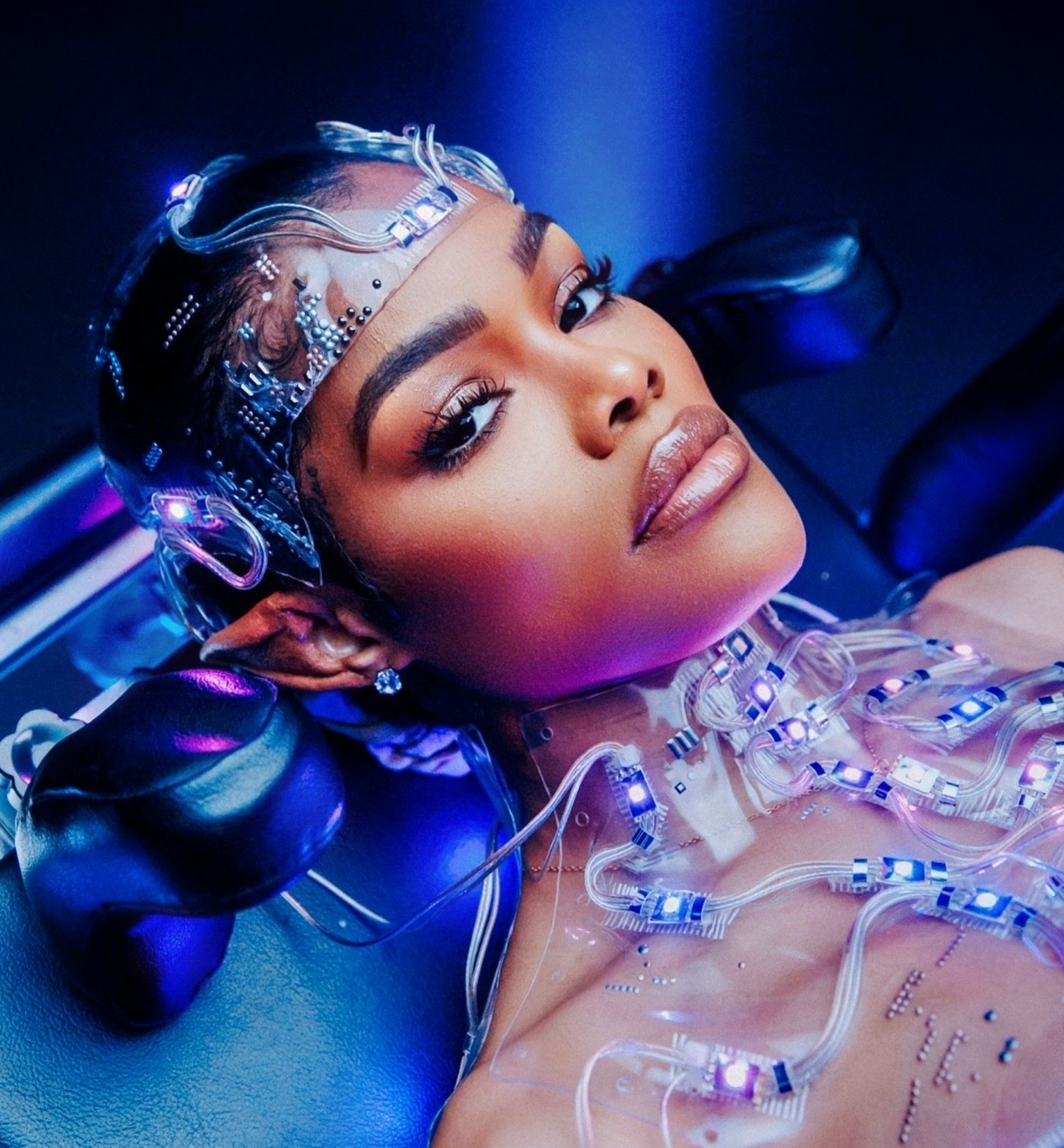 color photograph of an African American woman lying on a futuristic table surrounded by glowing blue and purple lights. She wears a transparent headpiece and chest apparatus embedded with small LED circuits and tubes.