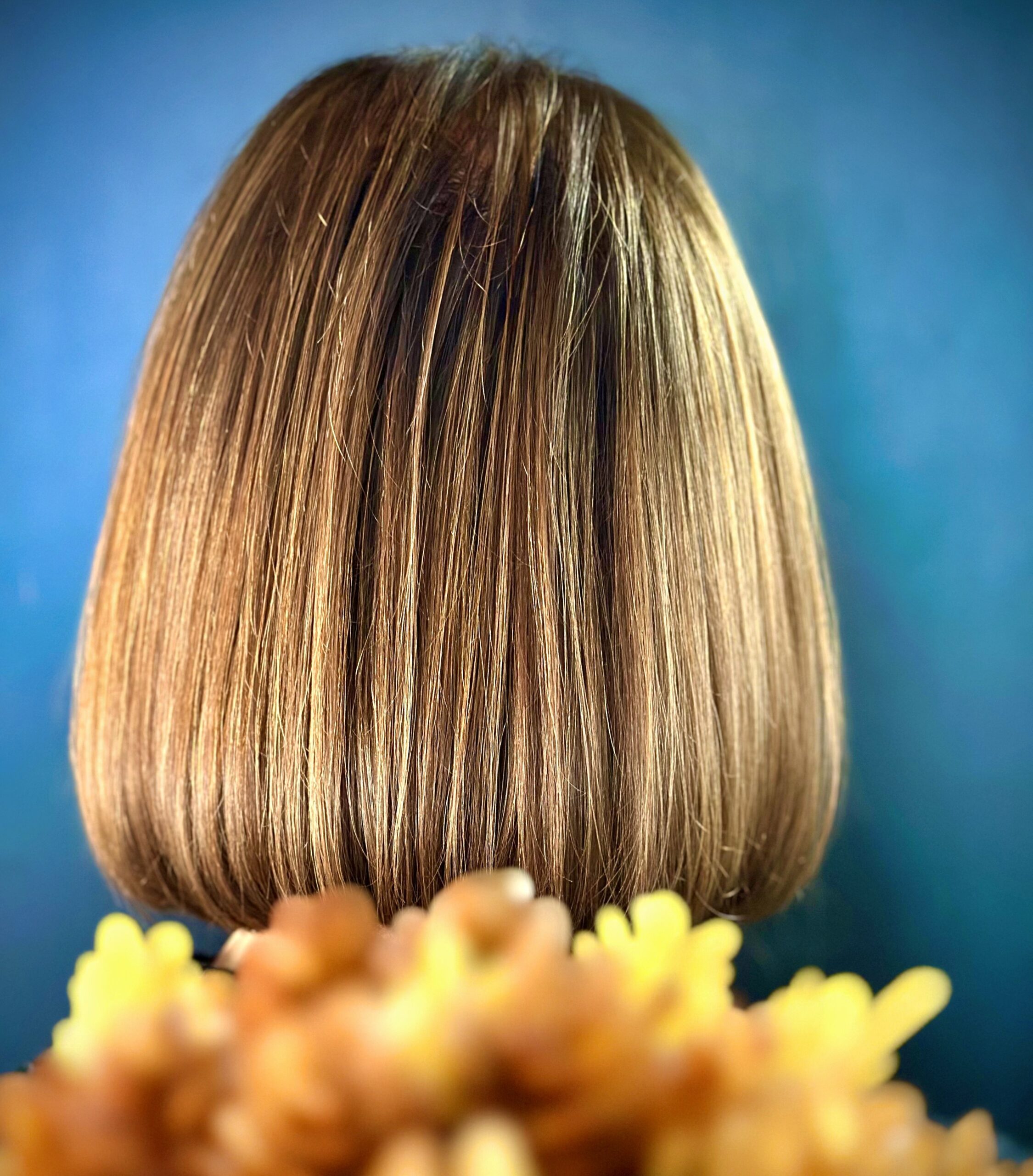A close-up color photograph of the back of a head with a bob hairstyle in front of a blue background with saffron yellow abstract foreground.
