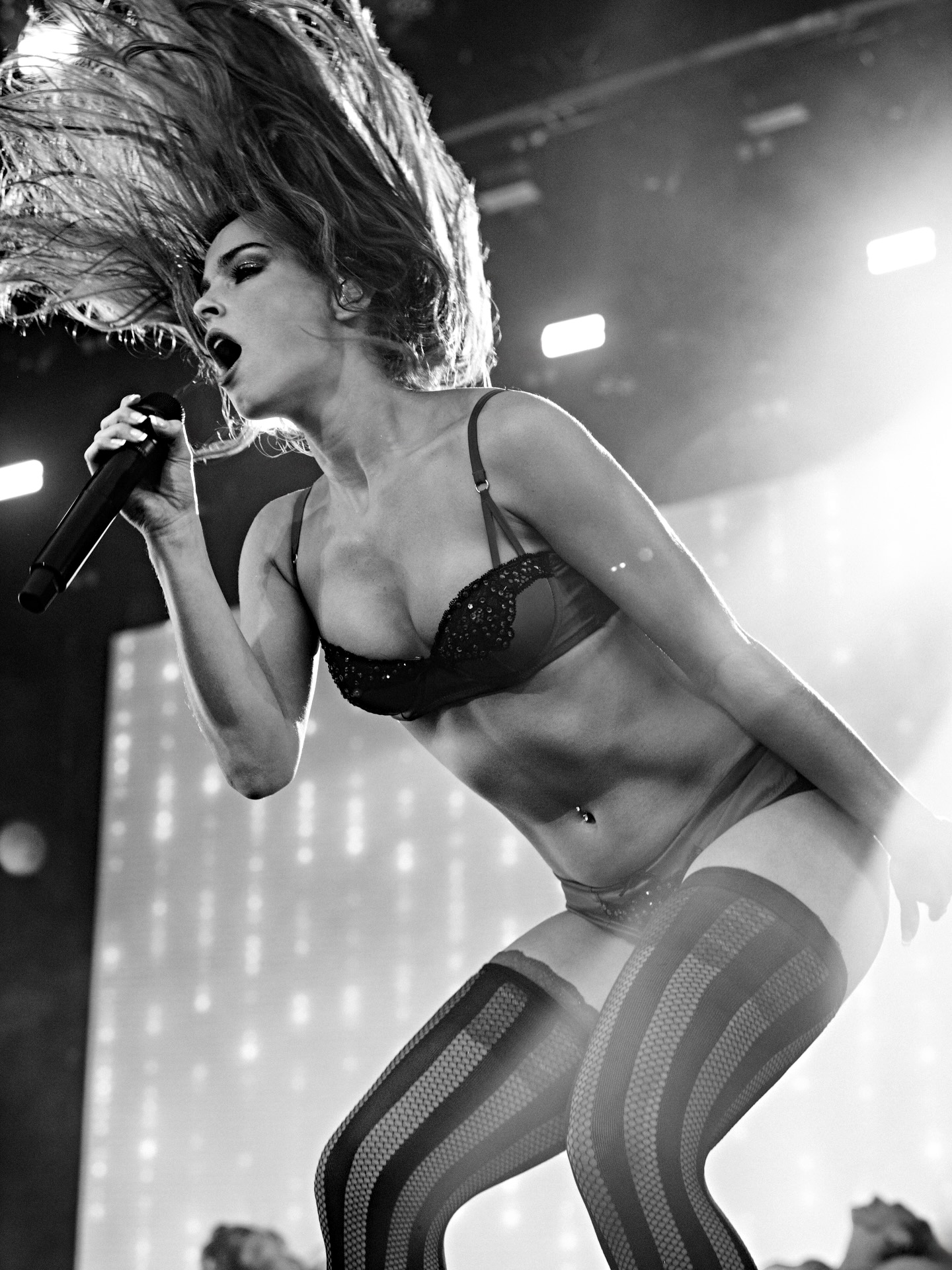 Black and white photograph of Addison Rae in a bra top and pants with thigh high stocking on a concert stage sining into a microphone