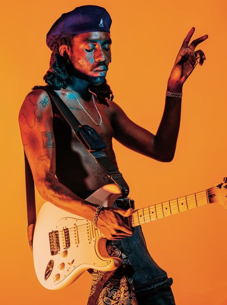 Color photograph of musician Dev Hynes on an orange background he is holding a guitar with his left hand and the left hand is raised up he is shirtless and has a beret on his head