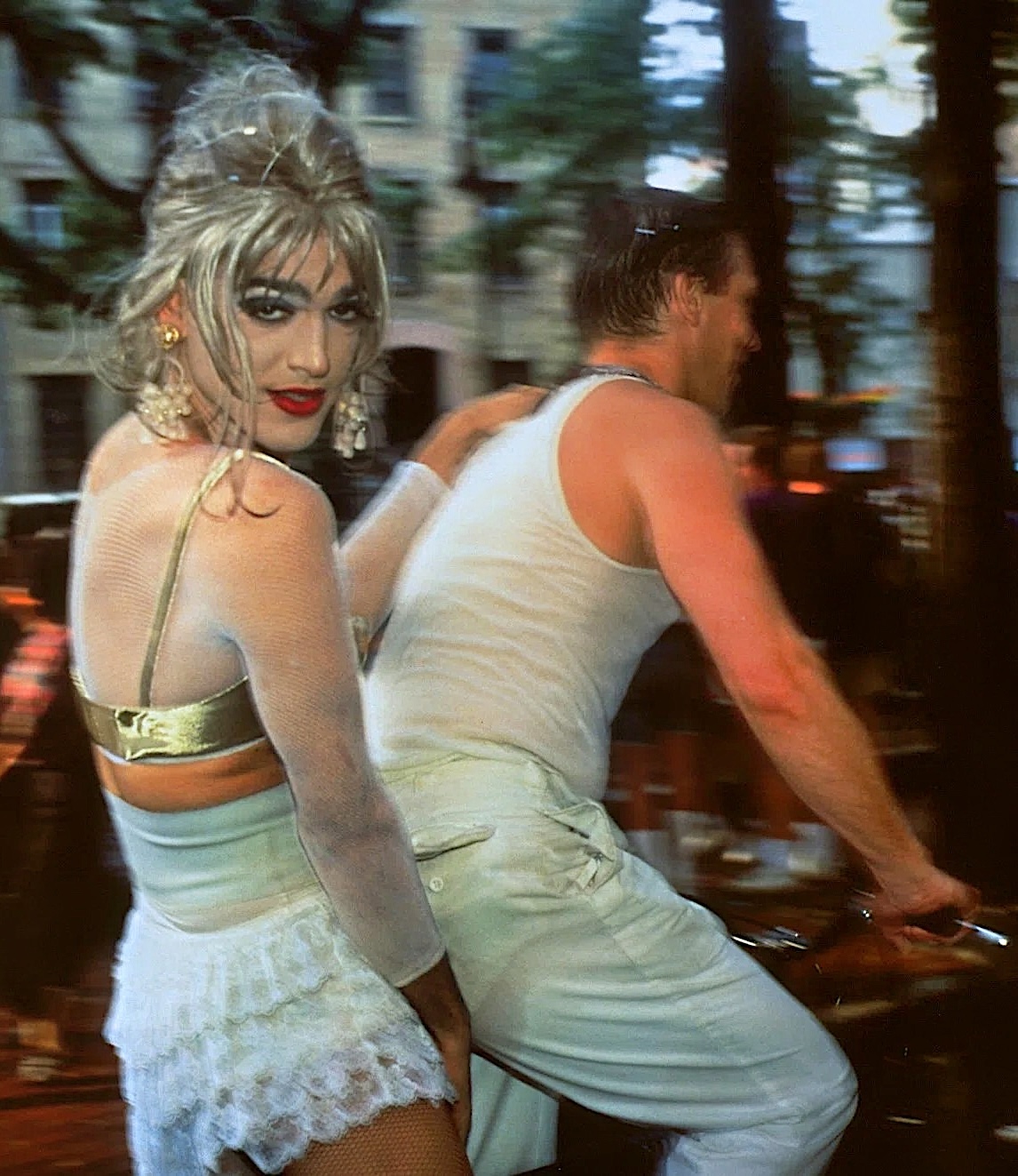 Color photograph of a queer man wearing makeup and a dress seated on the back seat of a bike with another man steering the bike