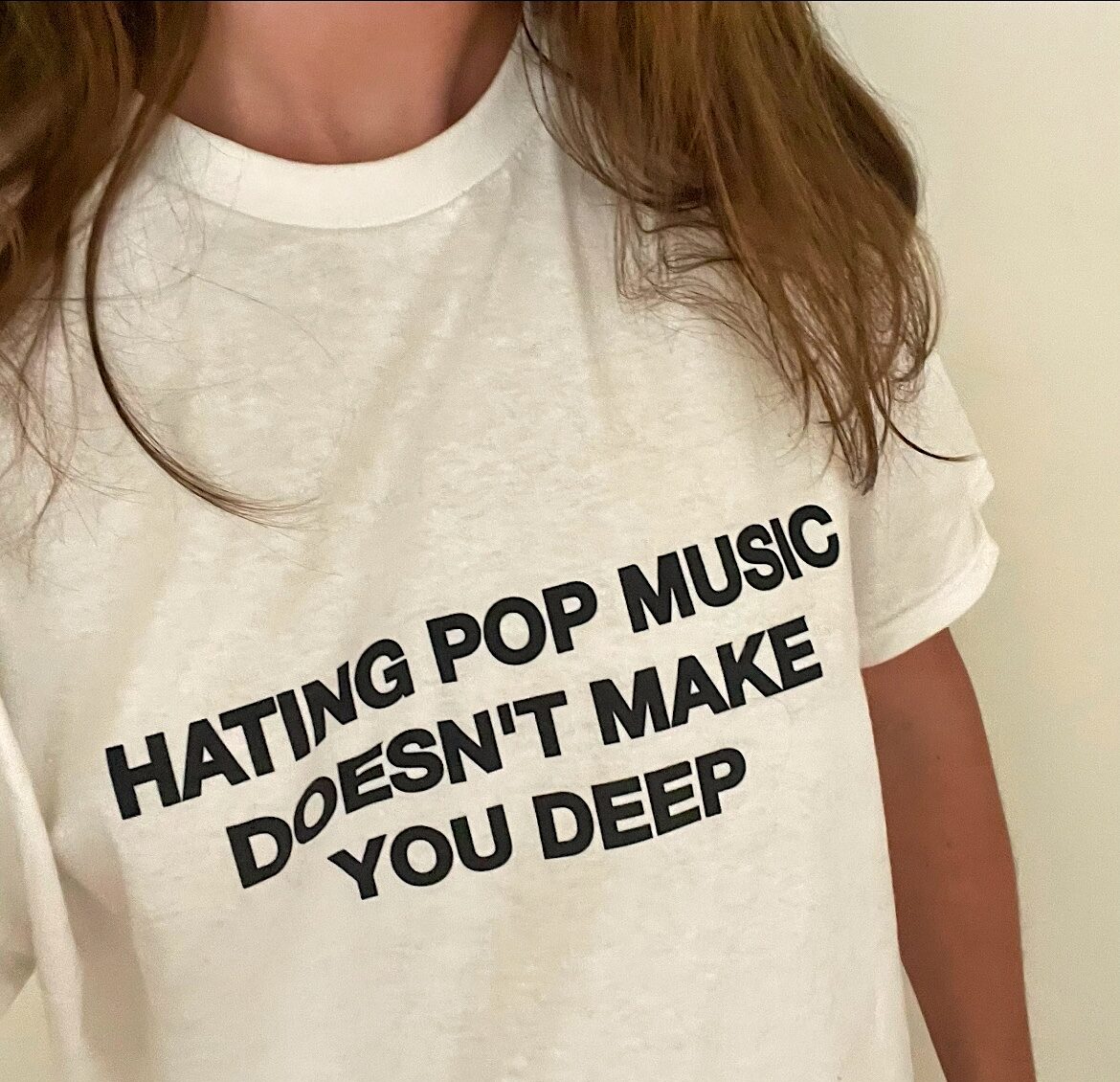 Close-up color shot of a person with long brown hair wearing a white t-shirt with bold black text that reads, "HATING POP MUSIC DOESN'T MAKE YOU DEEP."